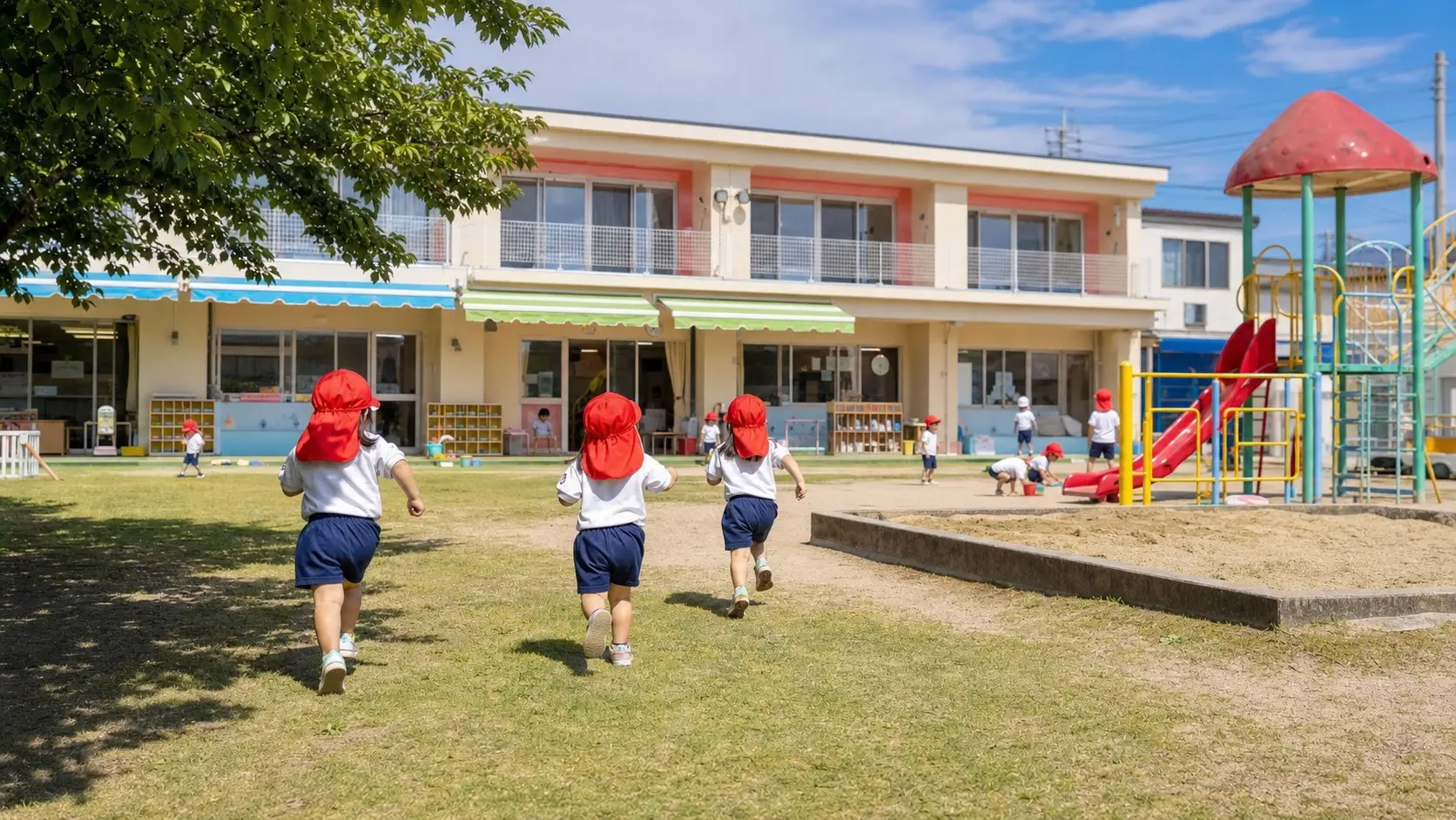 赤い帽子と体操着の子どもたちが園舎の前の園庭で遊ぶイメージカット
